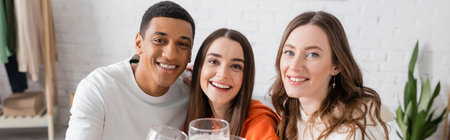 cheerful women and african american man looking at camera in living room, banner