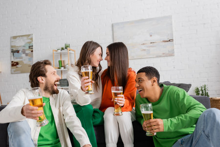 positive and interracial group of friends holding glasses of beer while looking at each other in living roomの写真素材