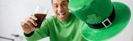 happy african american man holding glass of dark beer and green hat on Saint Patrick Day, banner