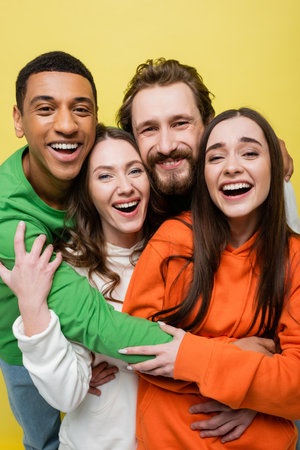 Happy interracial friends hugging and looking at camera isolated on yellowの写真素材