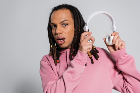 surprised multiracial man with dreadlocks holding wireless headphones on grey