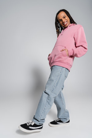 full length of happy multiracial man in blue jeans and pink hoodie posing with hands in pockets on...