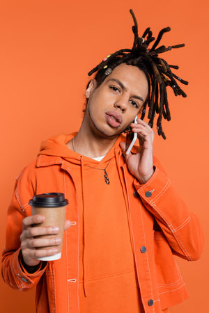 pierced and multiracial man with dreadlocks holding paper cup and talking on smartphone isolated on...