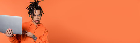 multiracial man with dreadlocks and tattoo holding laptop and looking at camera on coral background,...