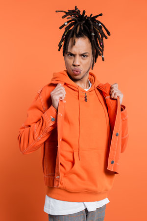 multiracial man with dreadlocks adjusting denim jacket while pouting lips on coral