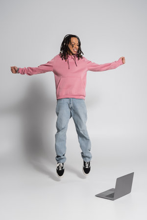 full length of happy multiracial man in pink hoodie and jeans jumping near laptop on grey