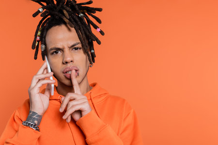 multiracial man with dreadlocks holding finger near lips and talking on cellphone isolated on coral