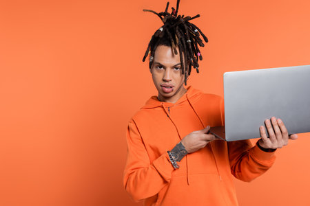 multiracial freelancer with dreadlocks and tattoo holding laptop and looking at camera on coral...