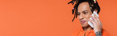 satisfied multiracial man with dreadlocks standing in hoodie and talking on smartphone isolated on coral, bannerの写真素材