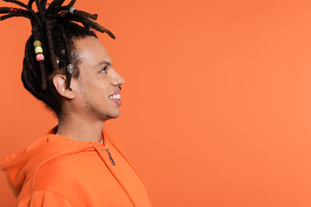side view of cheerful multiracial man with dreadlocks looking away and smiling isolated on coral...