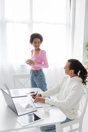 African american woman holding cup of coffee near boyfriend and gadgets at homeの写真素材
