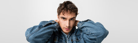 stylish young man in trendy denim clothes posing and looking at camera isolated on grey, banner