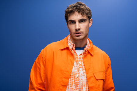 Young man in bright orange jacket standing isolated on blue