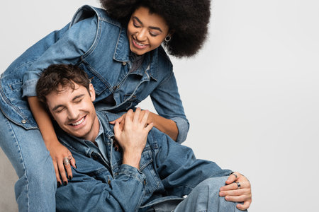 happy african american woman sitting and looking at cheerful man in denim outfit isolated on greyの写真素材