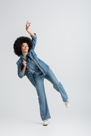 full length of curly african american woman in denim jacket looking at camera while posing with raised hand on greyの写真素材