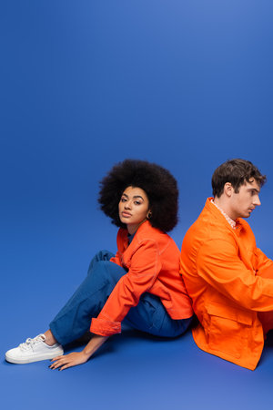 Curly african american woman in bright orange jacket sitting near man on blue backgroundの写真素材