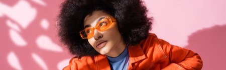 curly african american woman in stylish orange sunglasses looking away on pink, bannerの写真素材