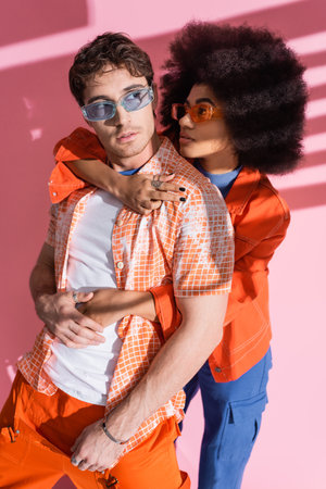 Trendy african american woman hugging man in orange overalls on pink background with shadow