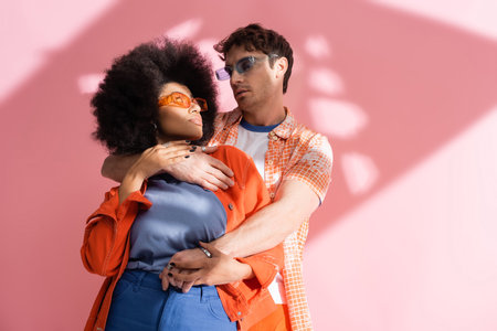 Man in sunglasses hugging stylish african american woman on pink background with shadow