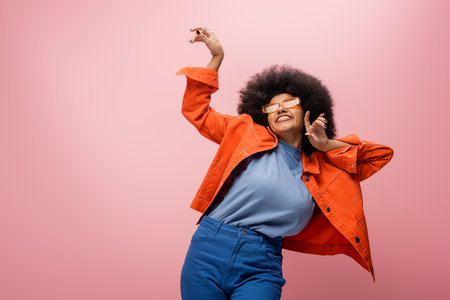 Cheerful african american model in sunglasses and orange jacket isolated on pink