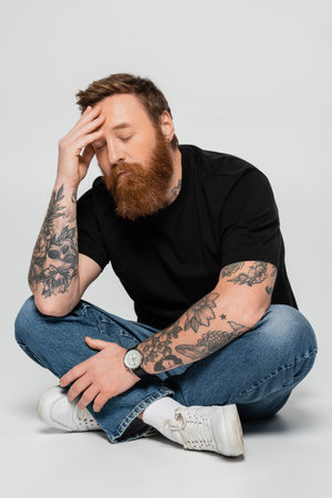 tired bearded man in jeans sitting with crossed legs and hand near head on grey backgroundの写真素材