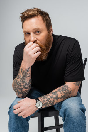 pensive tattooed man in black t-shirt and wristwatch holding hand near face and looking at camera isolated on greyの写真素材