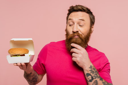 thoughtful tattooed man touching beard while looking at carton pack with tasty burger isolated on pinkの写真素材