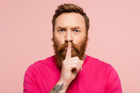 frowning bearded man showing hush sign and looking at camera isolated on pink