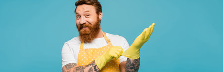 joyful bearded man in dotted apron putting on rubber gloves and smiling at camera isolated on blue, bannerの写真素材