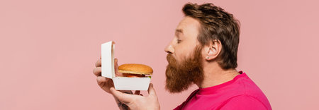 side view of bearded man smelling delicious hamburger isolated on pink, bannerの写真素材