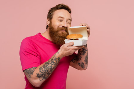 joyful tattooed man with closed eyes smelling tasty burger in carton box isolated on pinkの写真素材