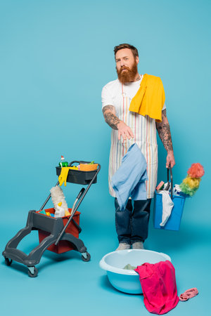 displeased man with garments and bucket pointing at laundry bowl near cart with cleaning supplies on blue backgroundの写真素材