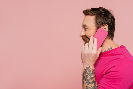 side view of cheerful man in magenta t-shirt calling on mobile phone isolated on pink