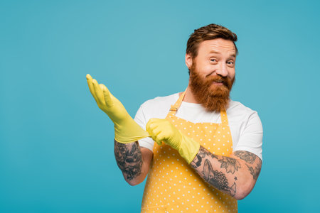 cheerful bearded man in apron looking at camera and wearing rubber gloves isolated on blueの写真素材