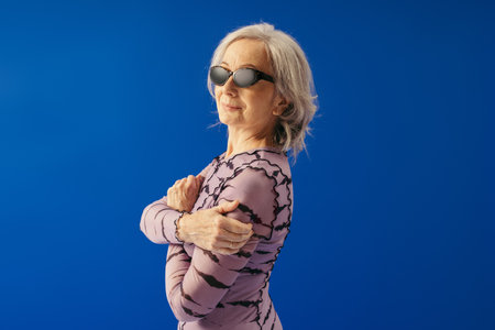cheerful senior woman in sunglasses and dress looking at camera isolated on blue