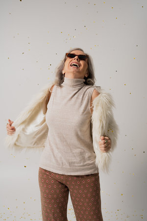 happy elderly woman in white faux fur jacket and sunglasses looking at falling confetti on grey...