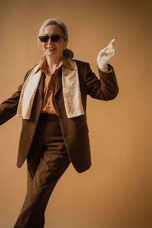 smiling senior woman in brown suit and white gloves gesturing on beigeの写真素材