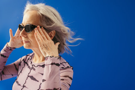 portrait of senior woman adjusting sunglasses while looking away isolated on blue