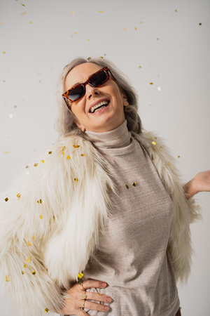 pleased elderly woman in white faux fur jacket and trendy sunglasses smiling near falling confetti...