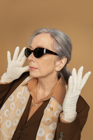 senior woman in white gloves and sunglasses posing isolated on beige