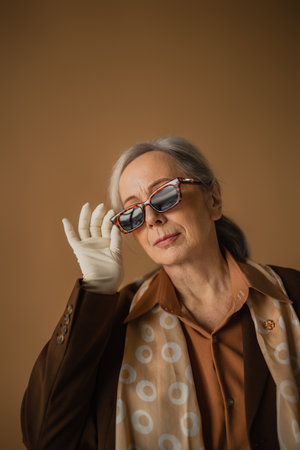 senior woman in brown suit and white gloves adjusting sunglasses on beige
