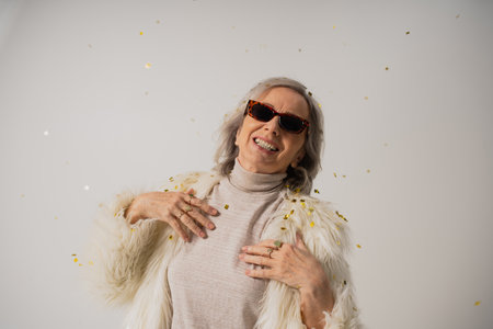 smiling senior woman in white faux fur jacket and trendy sunglasses near falling confetti on grey...