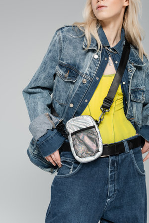 cropped view of blonde woman in stylish denim jacket with belt bag posing isolated on grey
