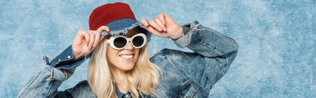 smiling blonde woman in denim jacket and sunglasses adjusting panama hat near blue textured...