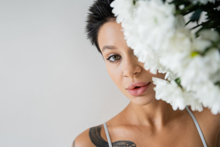 portrait of tattooed woman with makeup obscuring face with white flowers and looking at camera isolated on greyの写真素材
