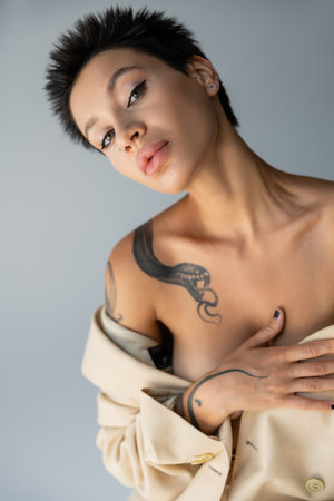 portrait of sexy tattooed woman with naked shoulders covering breast and looking at camera isolated on greyの写真素材