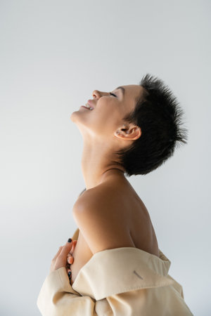 side view of happy and sexy woman in beige blazer with bare shoulders covering breast with hands isolated on greyの写真素材