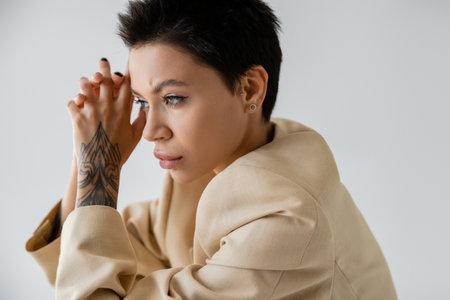 portrait of tattooed and thoughtful brunette woman in beige blazer holding hands near face isolated...