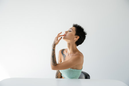 sexy tattooed woman touching lip while sitting near table and looking away on grey backgroundの写真素材