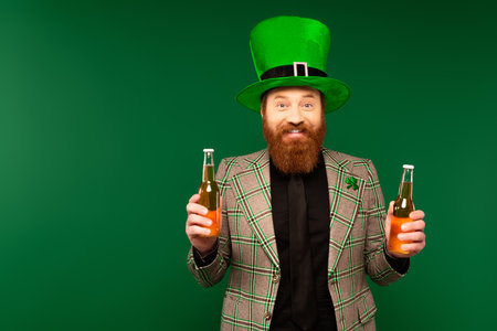 Smiling bearded man in hat holding bottles of beer isolated on green